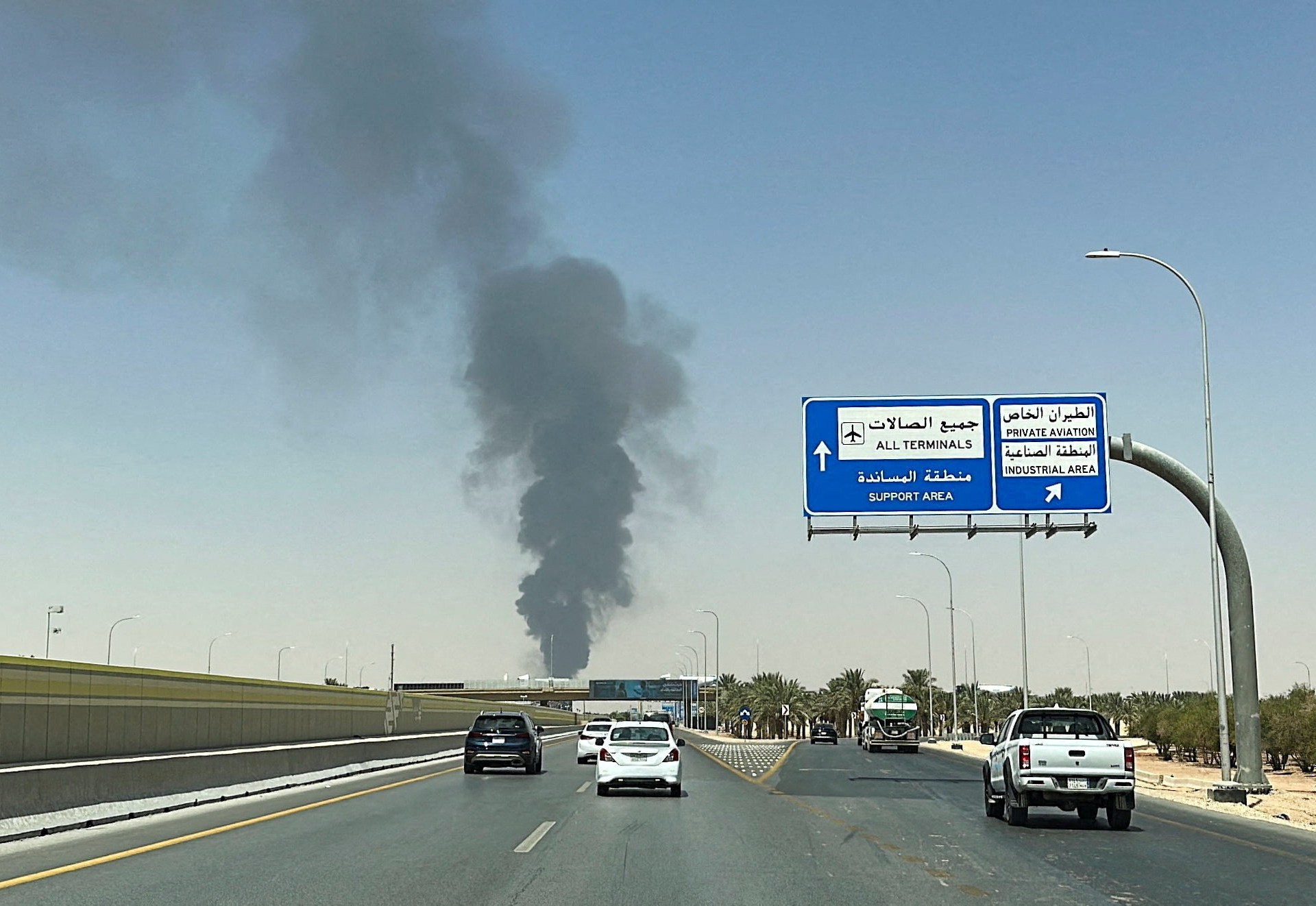 Thick black smoke plume beyond a Riyadh highway gantry pointing to King Khalid International Airport terminals — Stringer/Reuters via Al Jazeera